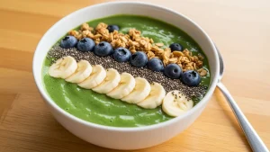 Vibrant green acai smoothie bowl topped with banana, blueberries, chia seeds, and granola on a wooden table.