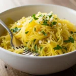 White bowl filled with golden spaghetti squash strands, bright green parsley, and minced garlic, with a fork, on a rustic wooden table.