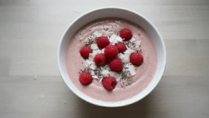 Thick, creamy low carb smoothie bowl with raspberries, shredded coconut, and chia seeds in a white bowl.