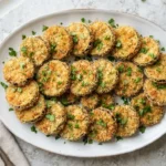 Golden-brown, crispy eggplant rounds on a white platter, garnished with green parsley, on a light grey surface.